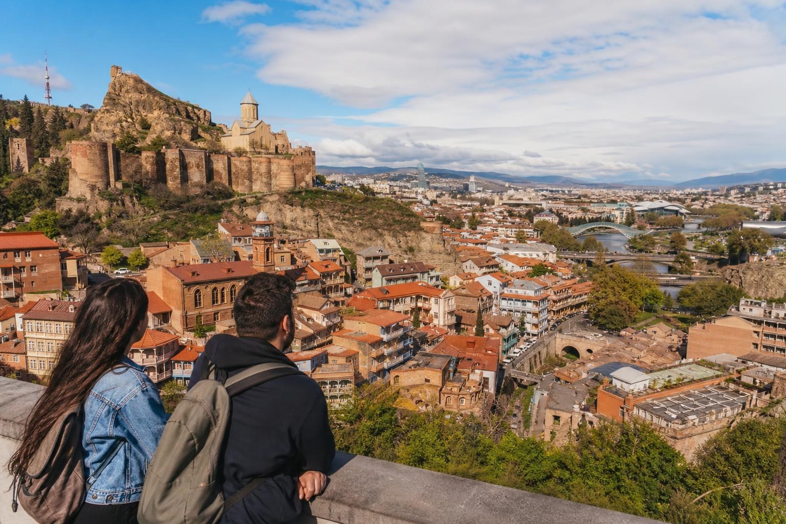 Two travelers overlook the Tbilisi skyline, featuring the Narikala Fortress, red-roofed houses, and the Bridge of Peace under a blue sky.