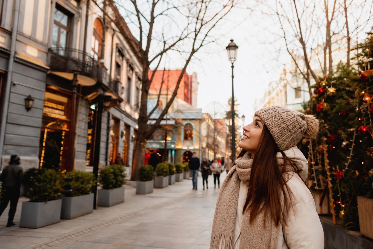 Smiling woman in winter clothes walking down a festive Tbilisi street lined with historic architecture and decorated Christmas trees.