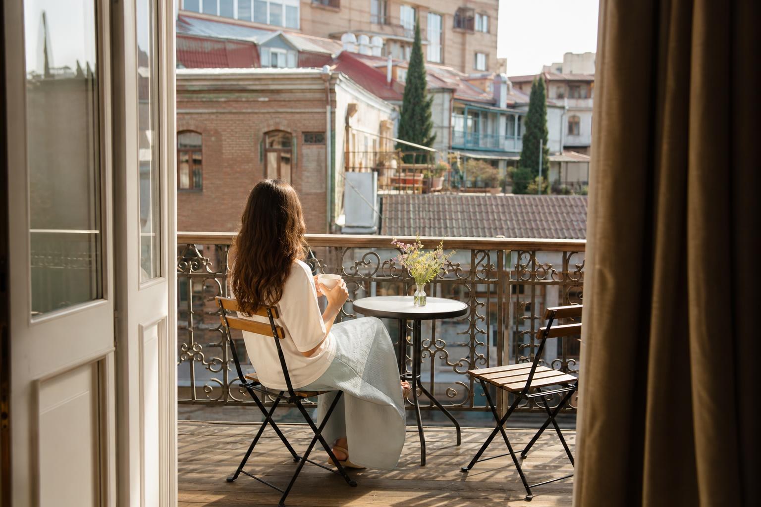 Woman sitting on a hotel balcony with a coffee cup, overlooking the historic brick buildings and rooftops of Tbilisi Old Town.