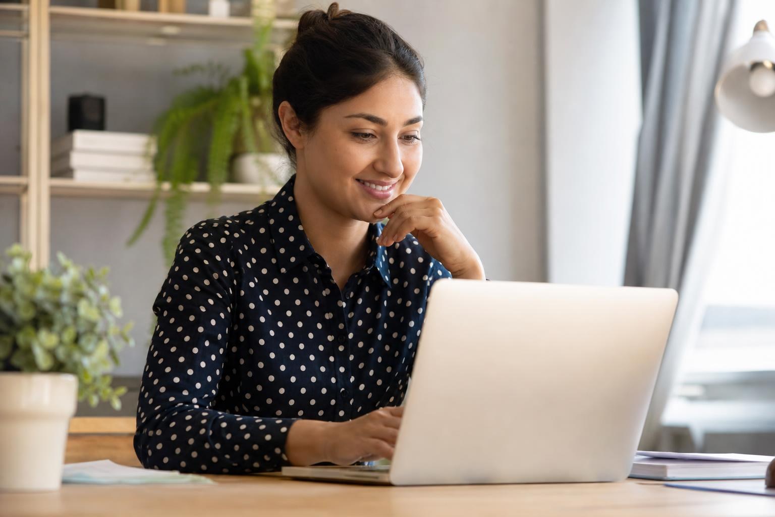 Smiling woman in a polka dot shirt using a laptop at a wooden desk in a bright, modern home office.
