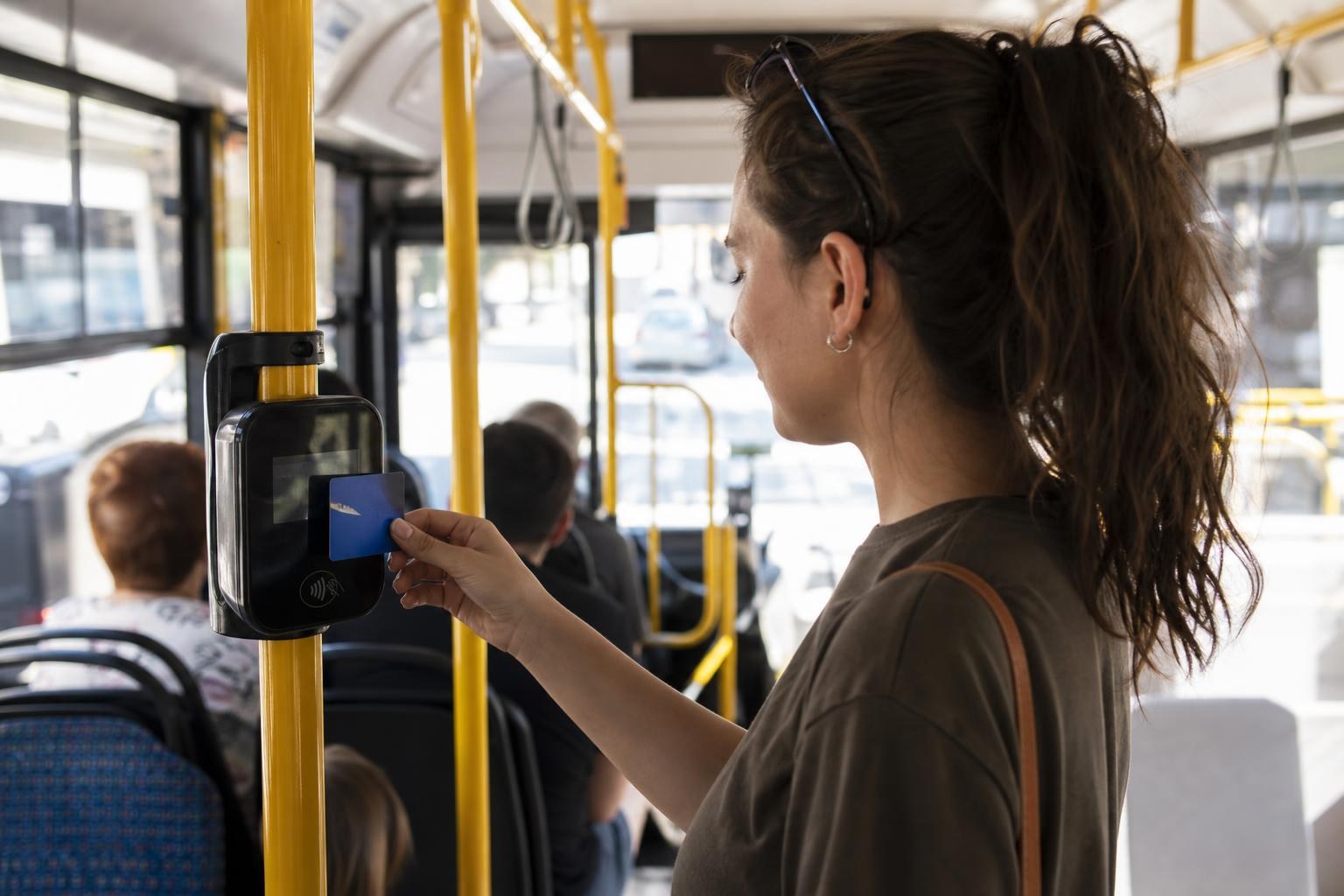 Woman tapping a blue transport card against a black electronic validator on a yellow pole inside a city bus.