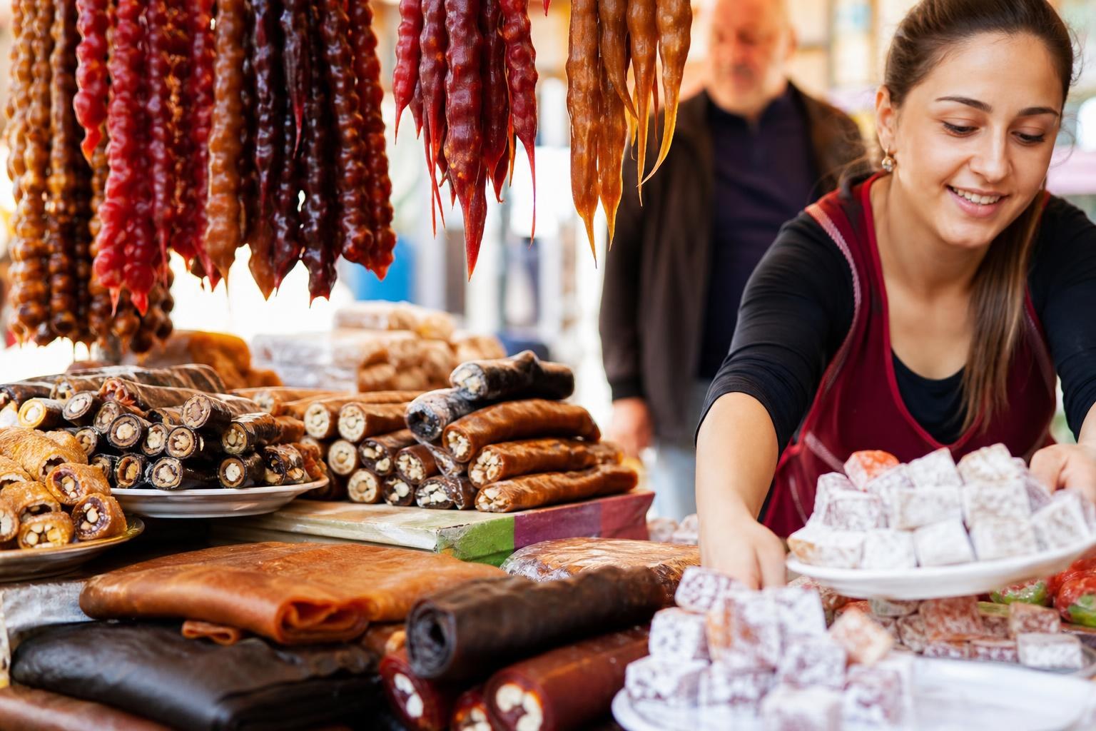 Smiling woman arranging plates of Turkish delight and churchkhela at a market stall filled with traditional Georgian sweets.