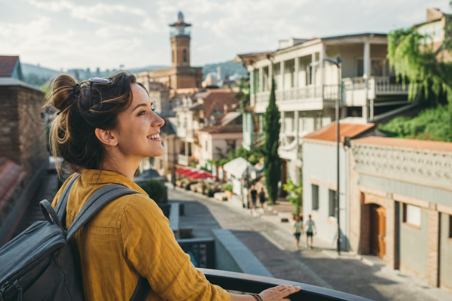 Smiling woman with a backpack overlooking the historic architecture and cobblestone streets of Old Tbilisi, Georgia.