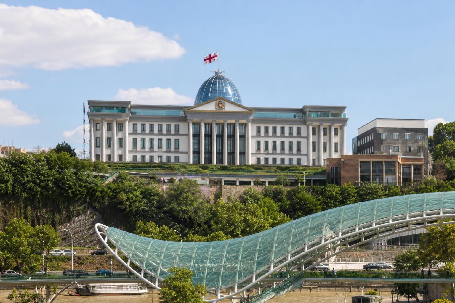 The Presidential Palace in Tbilisi with its glass dome and Georgian flag, viewed behind the modern glass canopy of the Bridge of Peace.