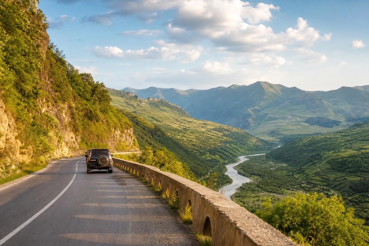 Black SUV driving on a winding mountain road overlooking a lush green valley with a winding river and distant peaks under a blue sky.