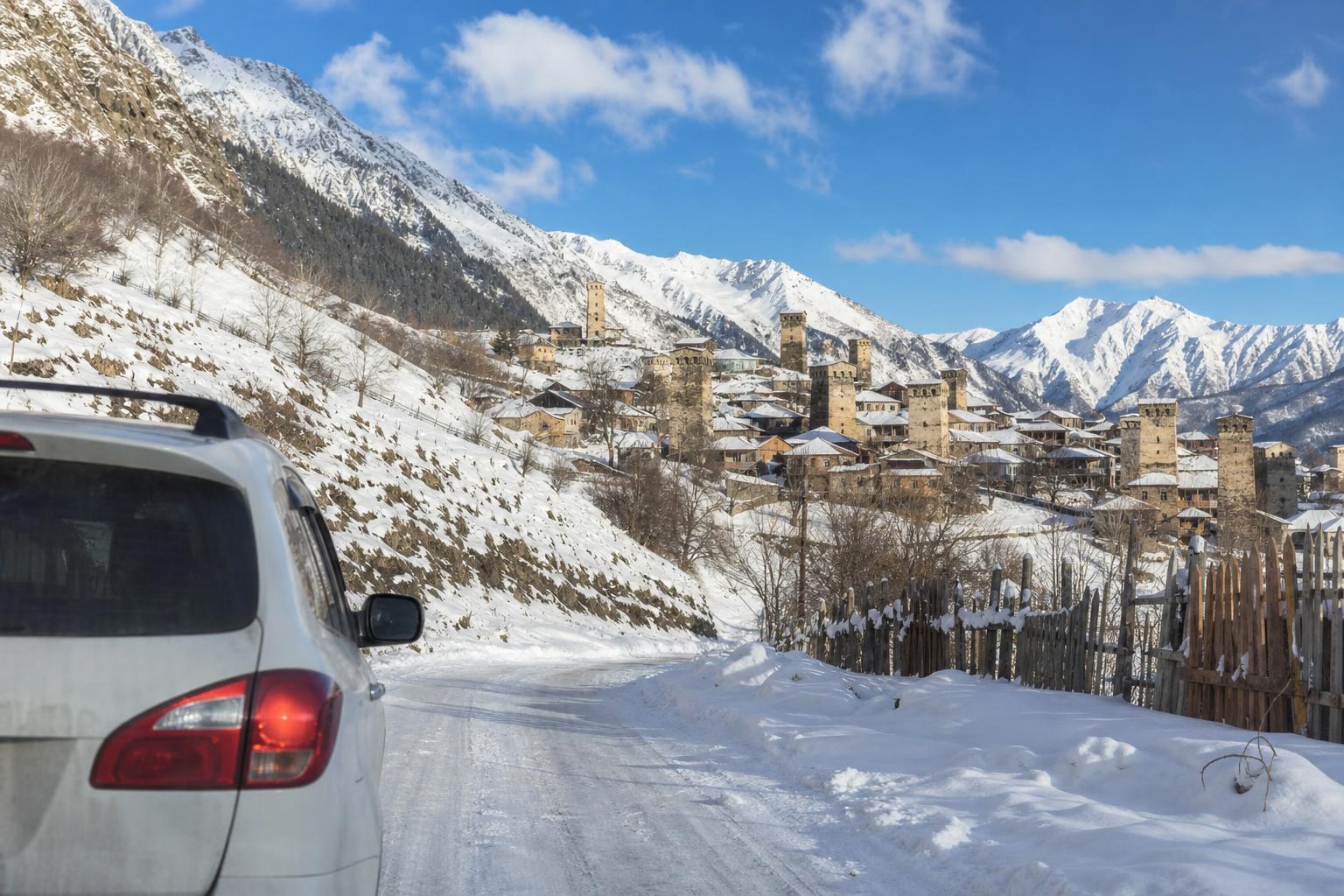 White SUV driving on a snowy mountain road toward a historic Svaneti village with stone defense towers under a bright blue sky.