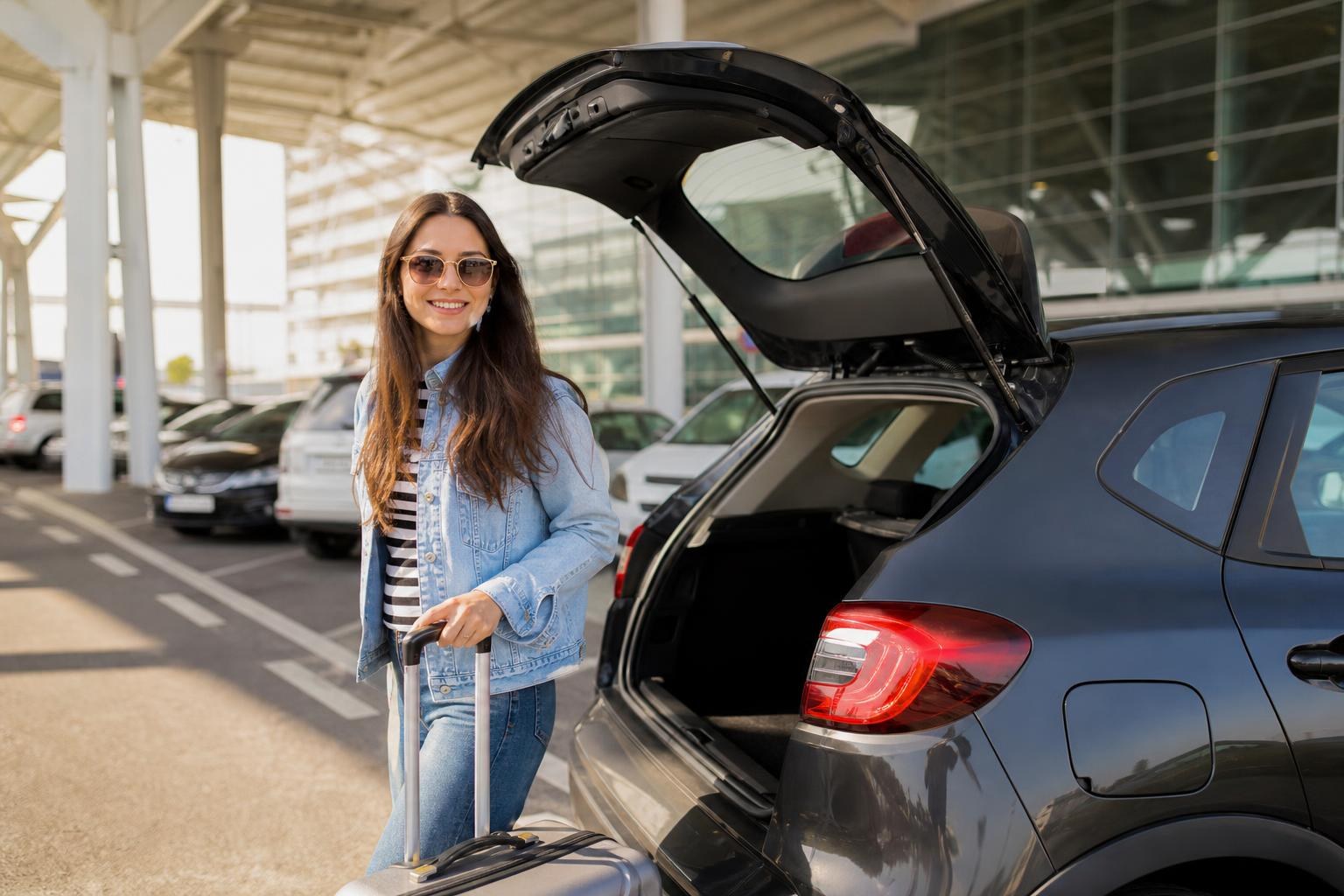 Smiling woman in a denim jacket standing with her suitcase next to the open trunk of a dark grey car at an airport parking lot.