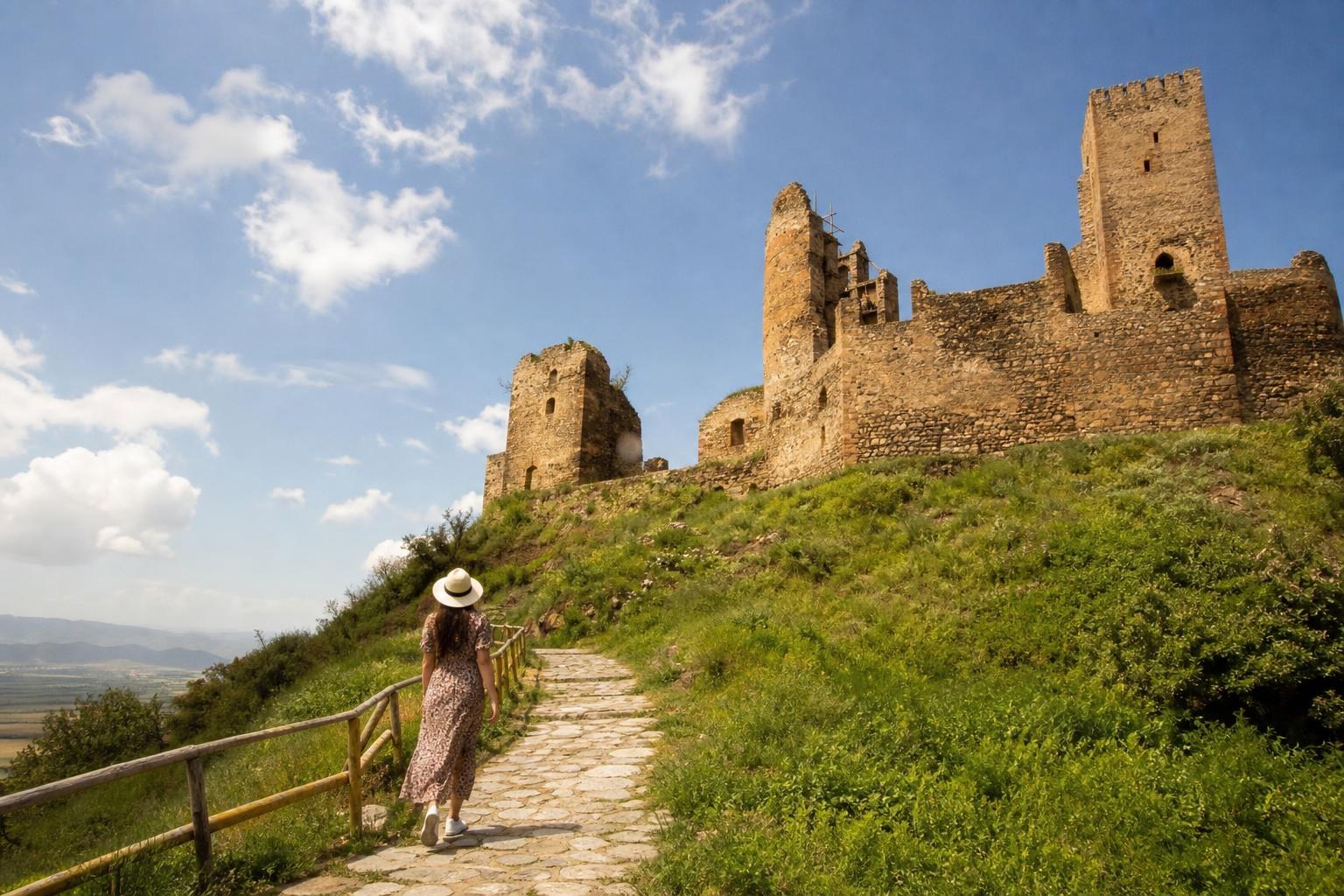 Woman in a sun hat walking up a stone path toward the ancient stone ruins of a hilltop fortress under a bright blue sky.